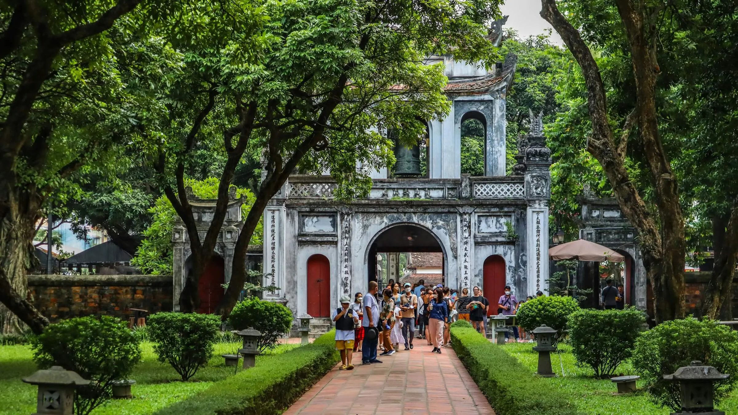 Van Mieu Gate, the stone entrance gate with a bell in its upper arch, at the end of a brick path between trees and hedges